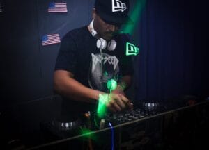 DJ performing in a nightclub with a sound mixer, wearing headphones and cap, under green and vivid lights.